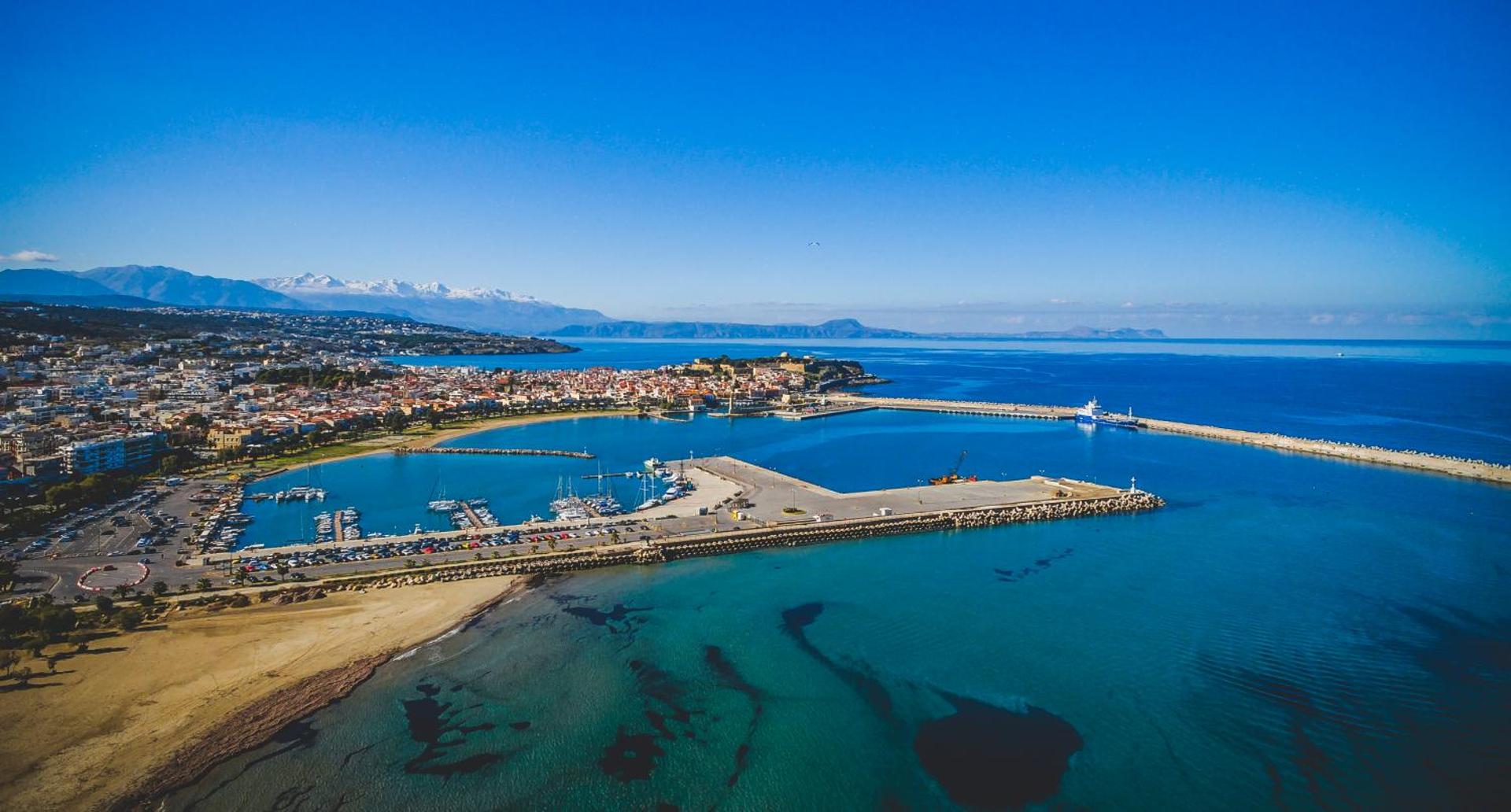 Dimitrios Beach viesnīcas apkārtne Retimno, Krētā — skats no augšas uz pilsētas ostu, zilo jūru un sniegotajām kalnu virsotnēm tālumā