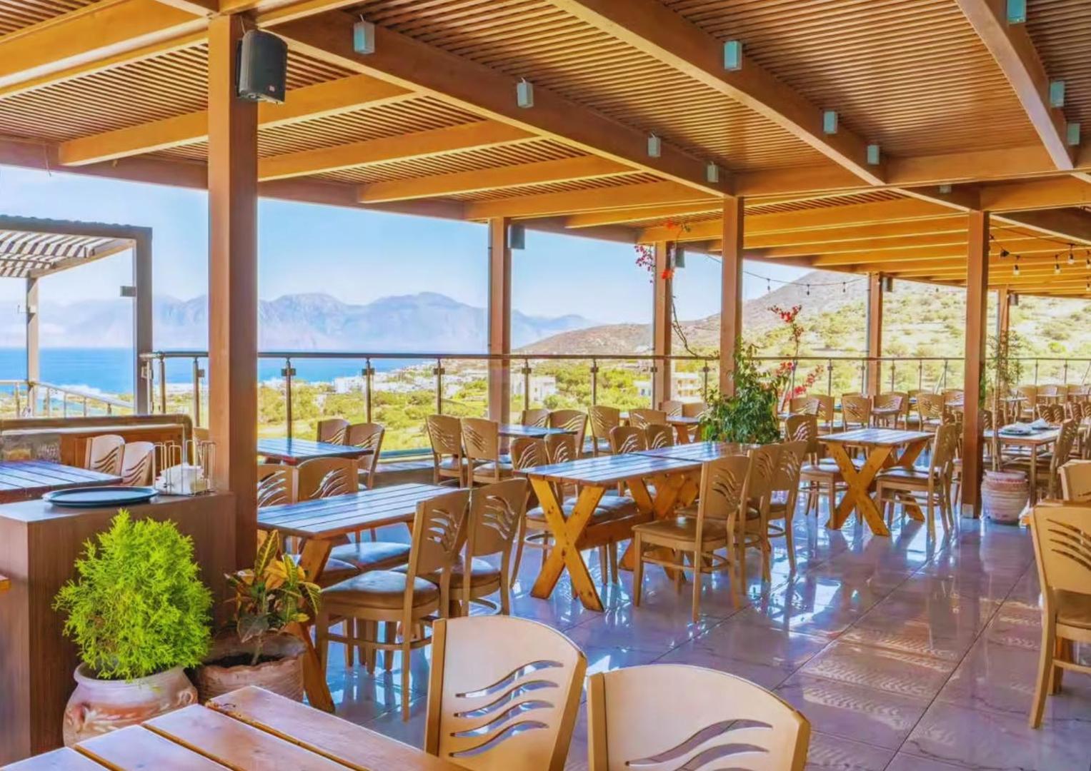 Elounda Waterpark Residence Hotel restorāna terase ar koka mēbelēm un panorāmas skatu uz Mirabello līci un kalniem Krētā