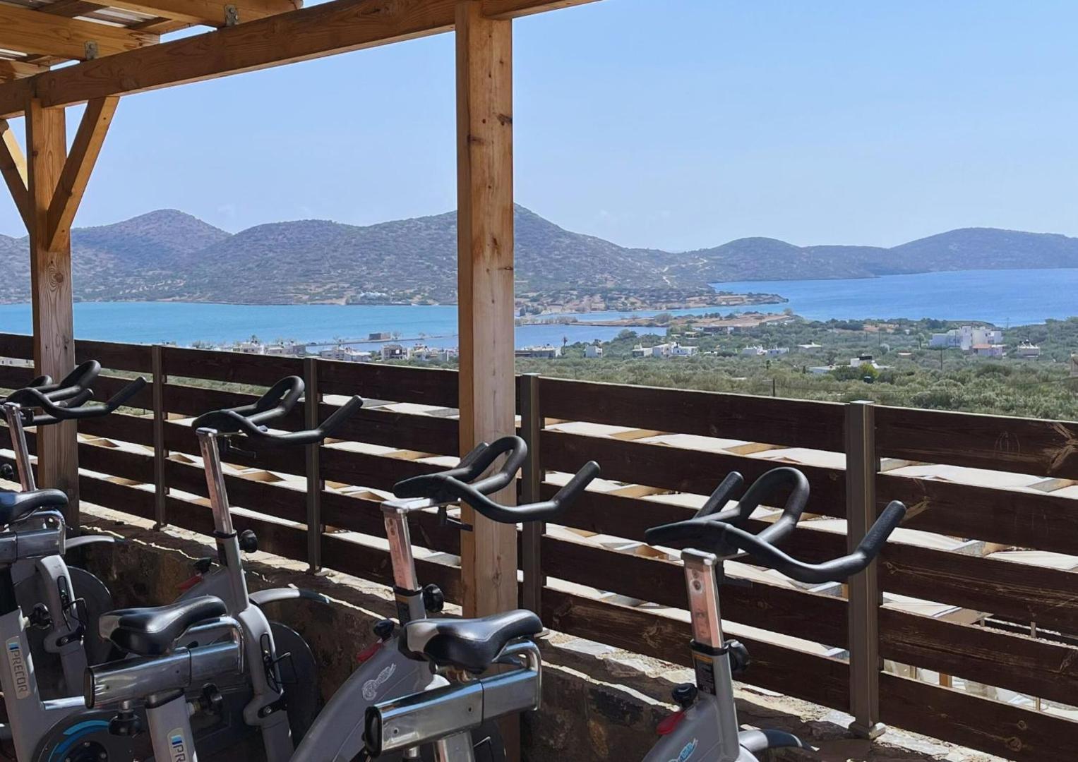 Elounda Waterpark Residence Hotel āra fitnesa zona ar velotrenažieriem un panorāmas skatu uz Elundas līci un kalniem