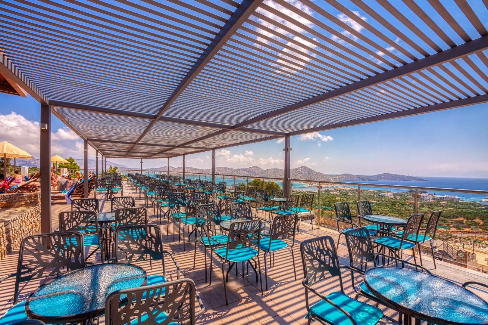 Elounda Waterpark Residence Hotel plašā āra terase ar moderniem galdiņiem un ziliem krēsliem zem saules aizsega Elundā, Krētā