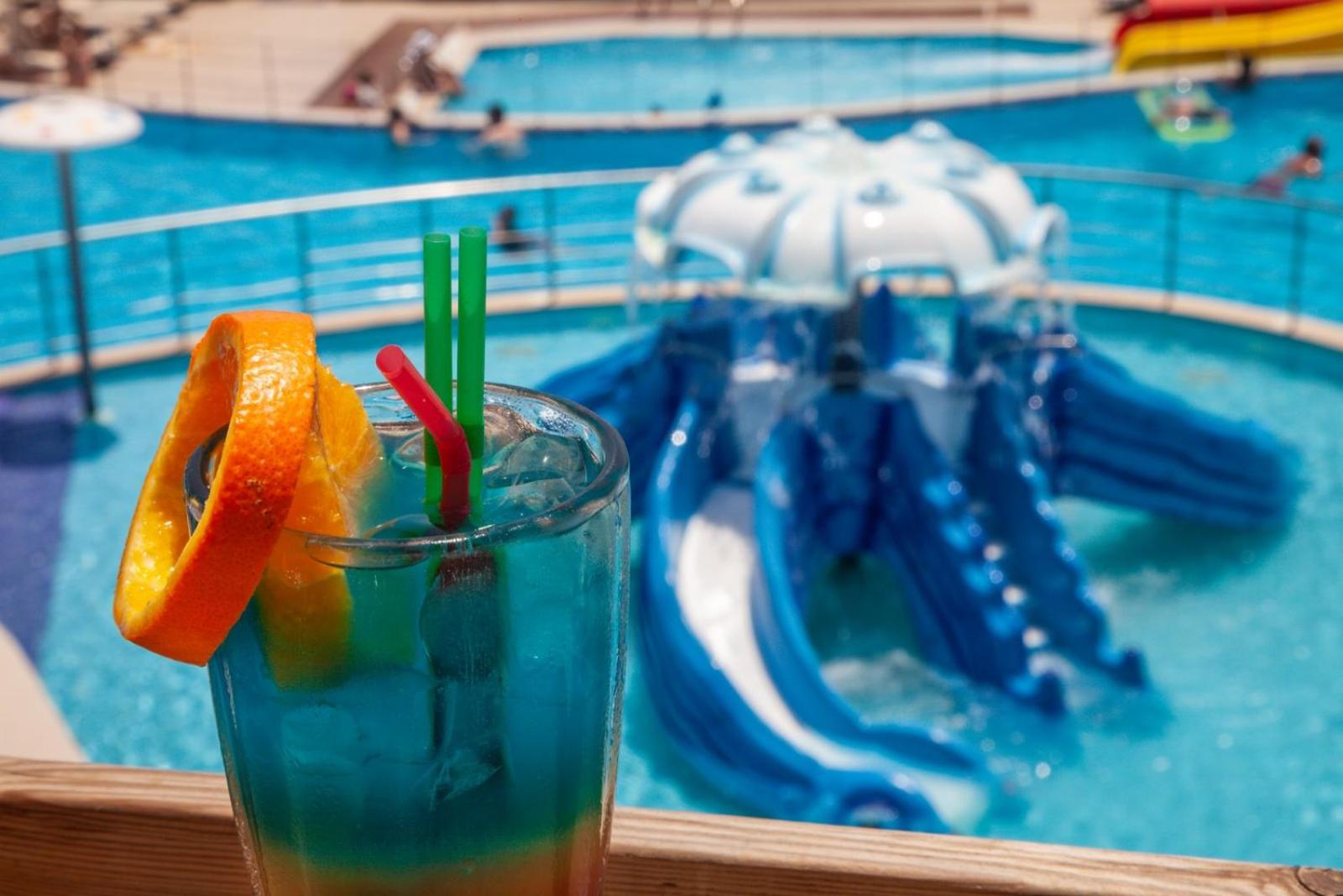 Skats uz Elounda Waterpark Residence Hotel akvaparku un zilo baseinu caur krāsainu kokteili ar apelsīna šķēli
