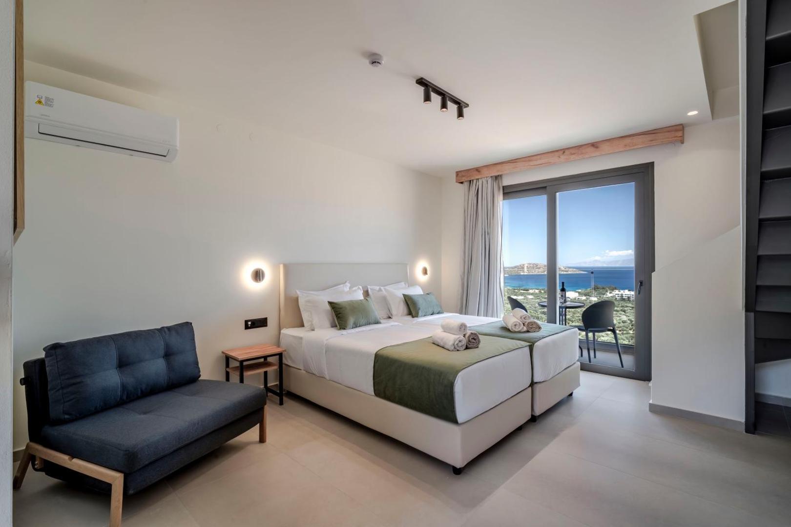 Elounda Waterpark Residence Hotel moderns divvietīgs numurs ar balkonu, skatu uz jūru, pelēku dīvānu un gaišu interjeru Krētā