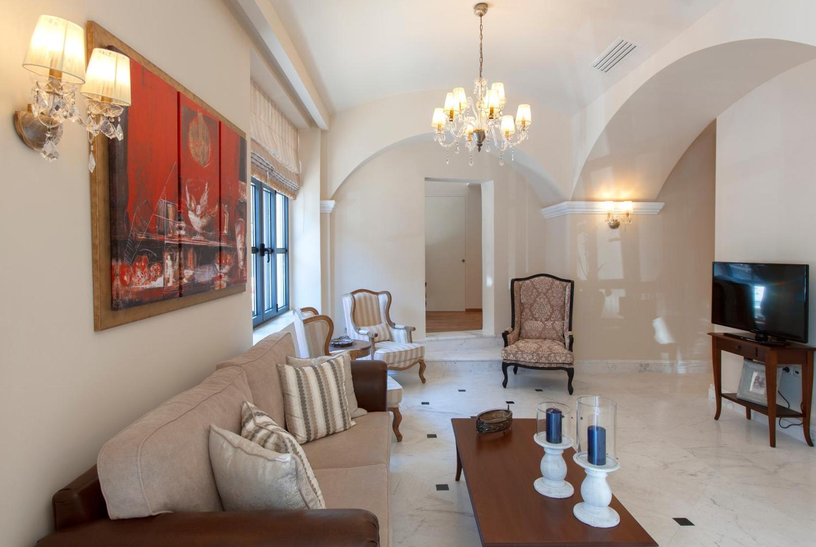 Ionia Suites viesnīcas numura atpūtas zona ar gaišu dīvānu, klasiskiem atpūtas krēsliem, marmora grīdu un elegantu lustru