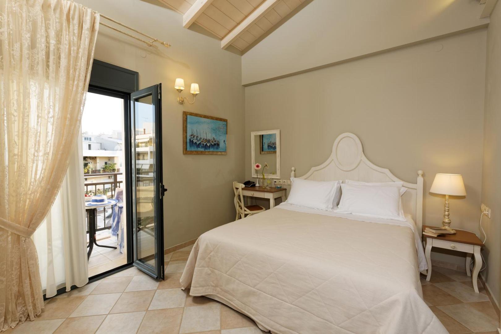Ionia Suites mājīgs numurs ar divguļamo gultu, gaišām mēbelēm un izeju uz balkonu, no kura redzama pilsētas ainava