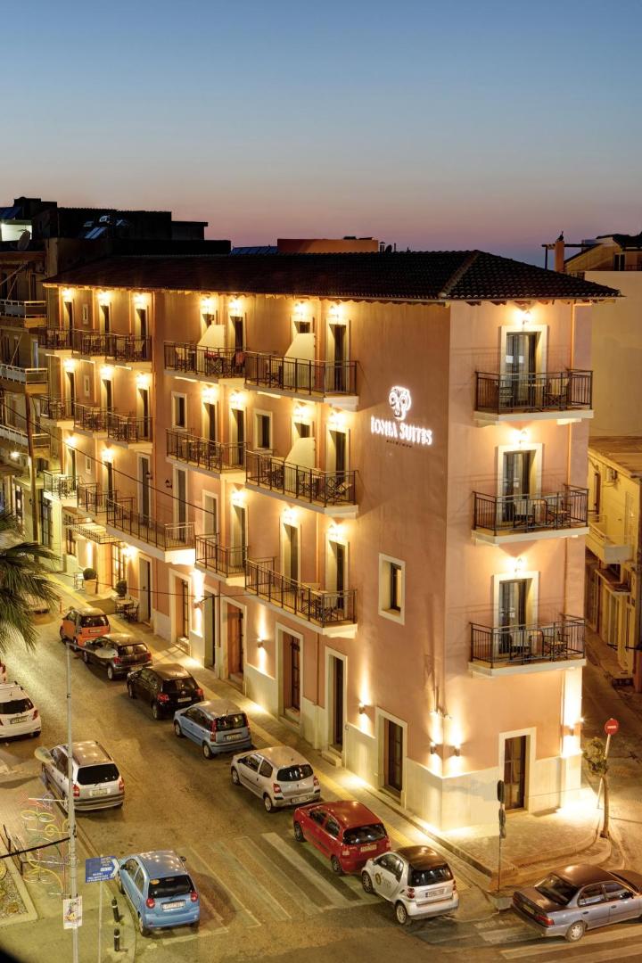 Ionia Suites viesnīcas fasāde vakarā Retimno pilsētā Krētā — četru stāvu ēka ar balkoniem un izgaismotu logo pie pilsētas ielas