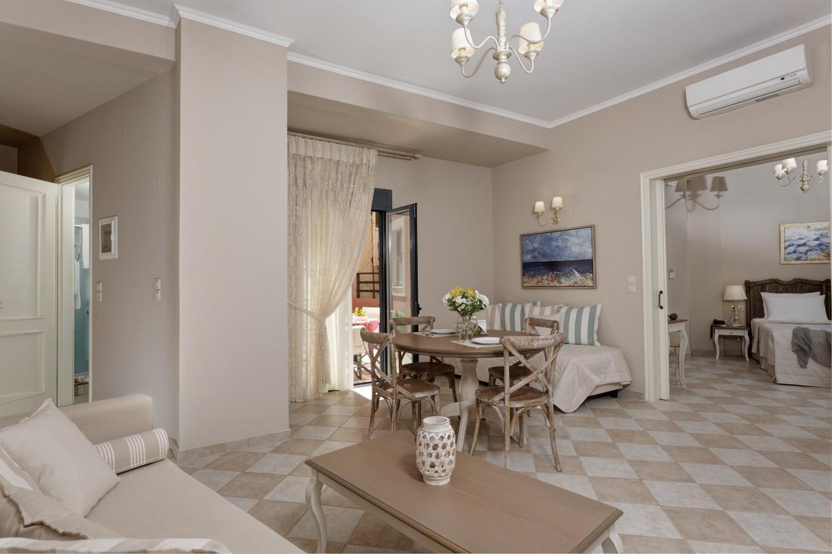 Ionia Suites plašs numurs ar viesistabas zonu, pusdienu galdu, dīvānu un atvērtām durvīm uz guļamistabu gaišos toņos