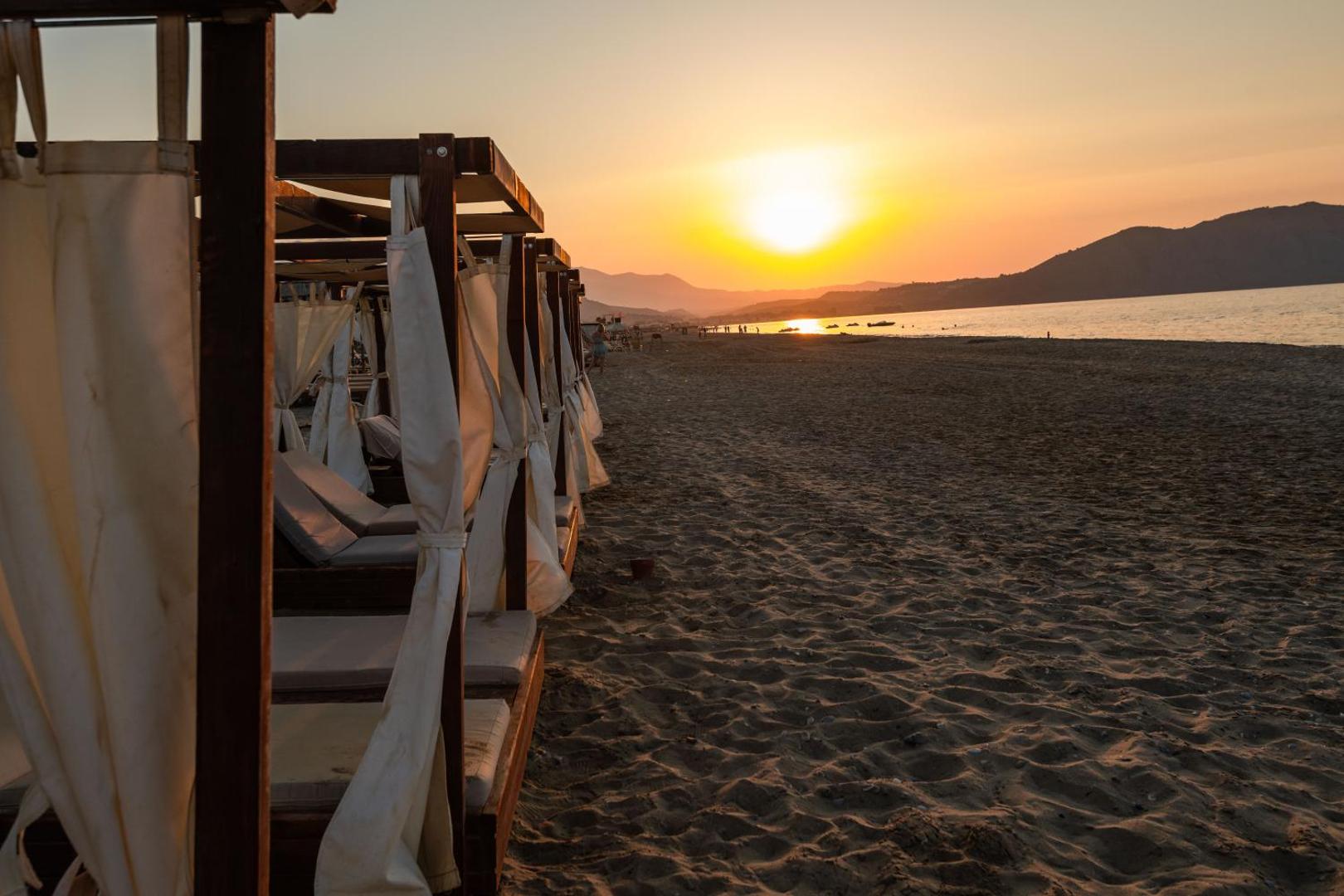 Ekskluzīvas pludmales teltis ar aizkariem Happy Days Beach kūrortā saulrieta laikā, piedāvājot privātumu un komfortu pie jūras.
