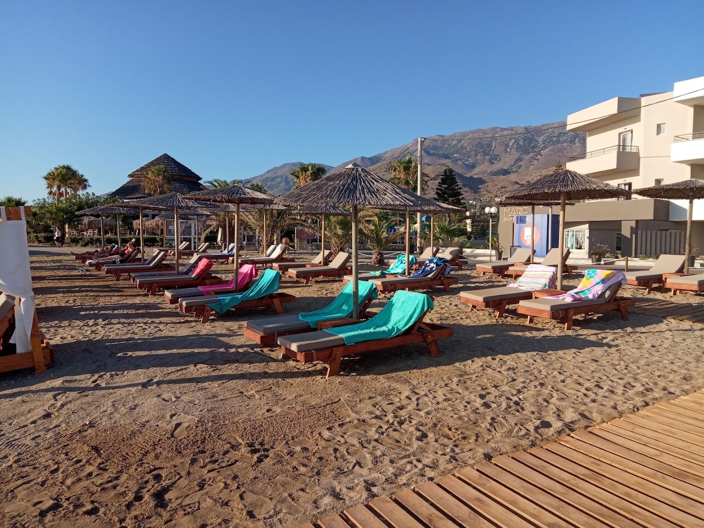 Mare Blue Suites viesnīcas pludmales zona ar saulessargiem, zviļņiem un skatu uz kalniem Stalidā, Krētas salā.