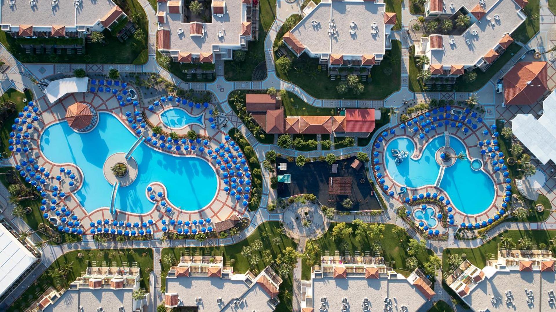 Lindos Princess Beach Hotel baseinu komplekss no augšas ar atpūtas krēsliem, bērnu zonu un apkārtējiem bungalo numuriem.