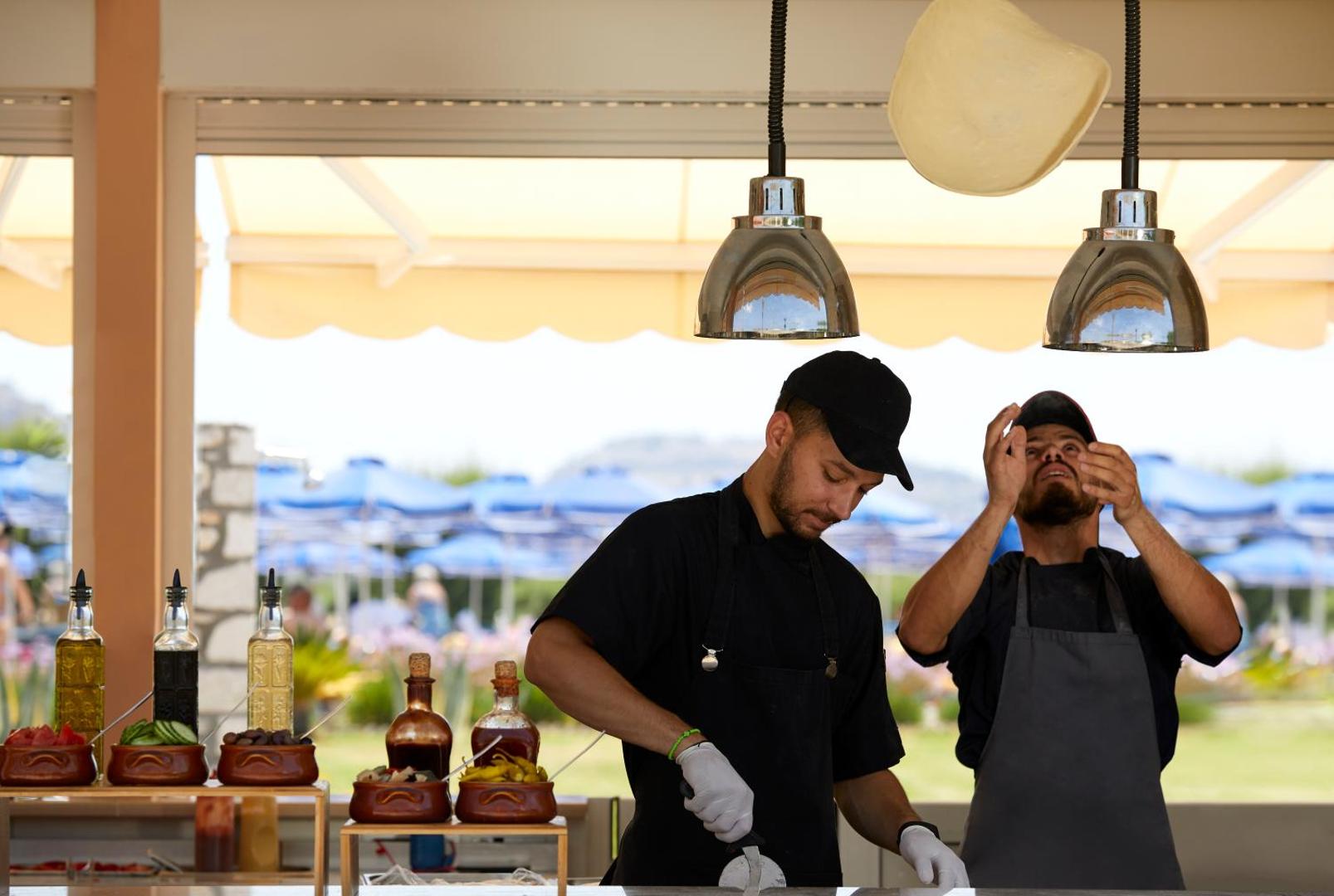 Lindos Princess Beach Hotel pavāri gatavo picu brīvdabas restorānā Rodas salā — redzama mīklas mešana un svaigas piedevas uz letes