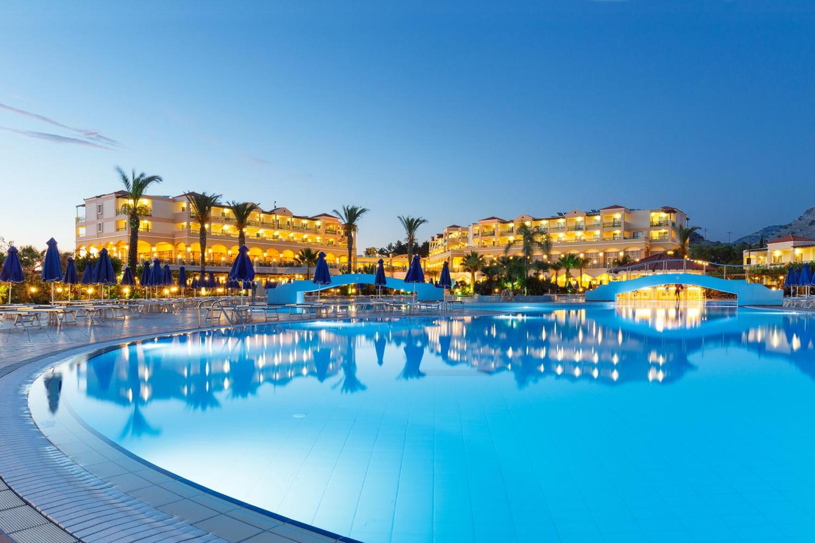 Lindos Princess Beach Hotel galvenais baseins vakarā Rodas salā — izgaismota viesnīcas fasāde, zili saulessargi un palmas pie ūdens