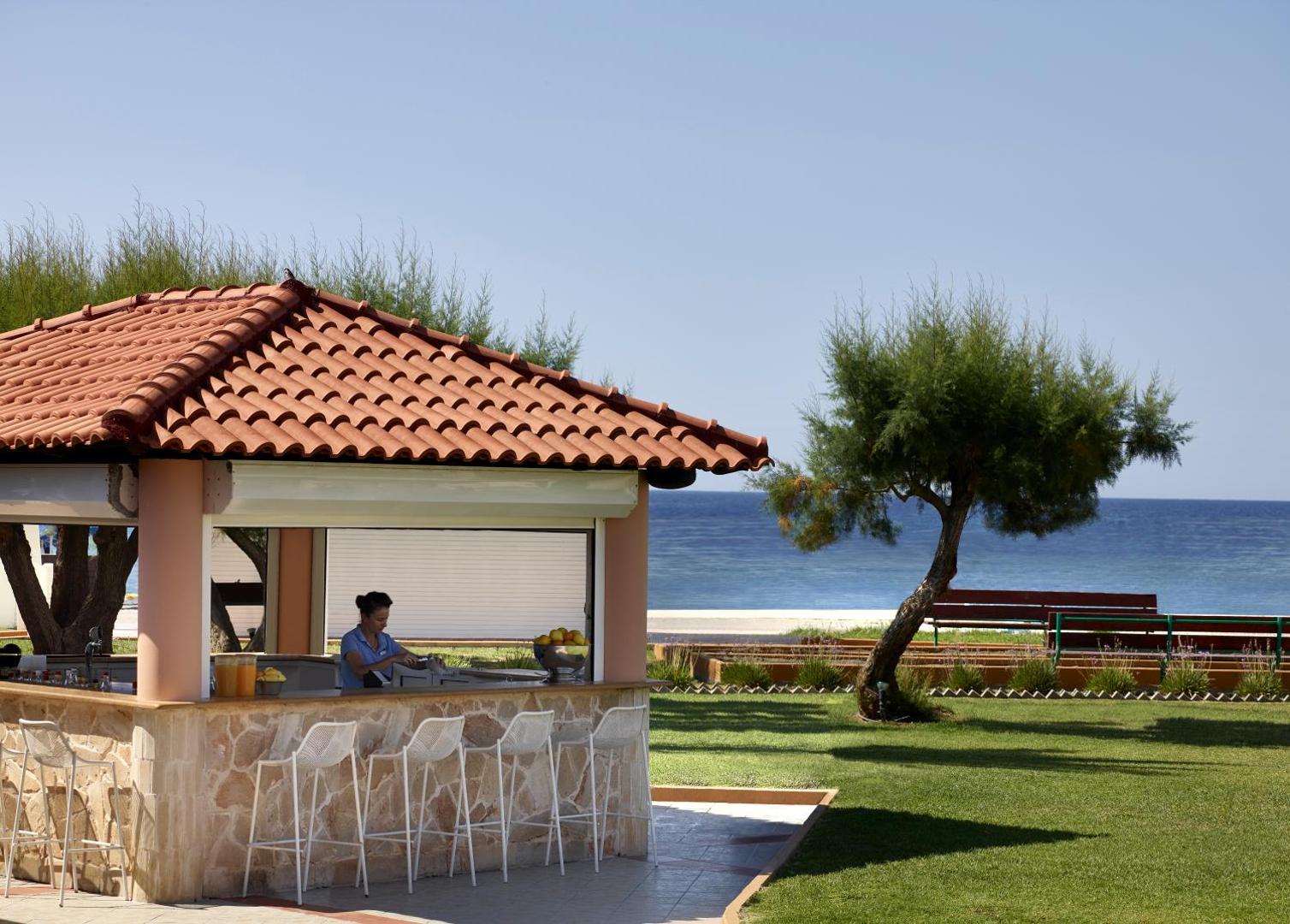 Lindos Princess Beach Hotel pludmales bārs ar akmens apdari un dakstiņu jumtu Rodas salā, Grieķijā — skats uz zilo jūru un zālāju