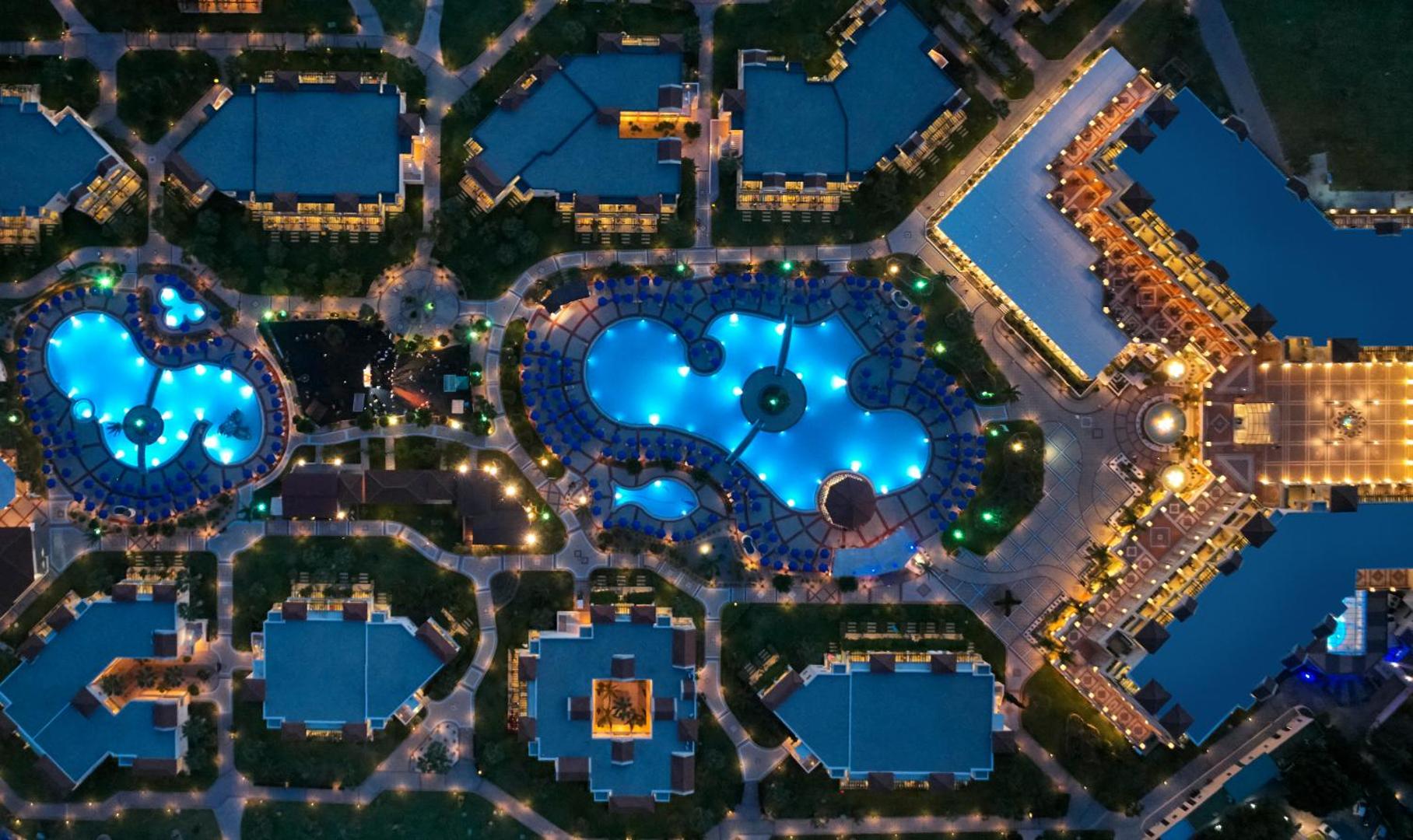 Lindos Princess Beach Hotel naksnīgs skats no putna lidojuma uz izgaismotiem baseiniem un bungalo korpusiem Rodas salā, Grieķijā.