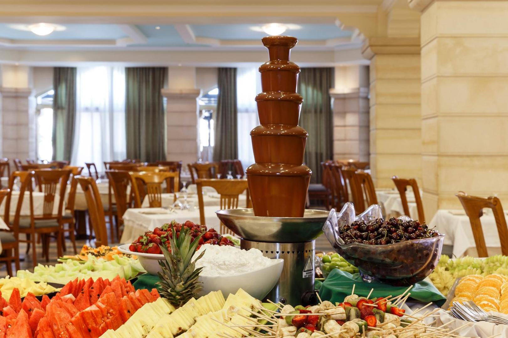 Lindos Princess Beach Hotel bufetes restorāna deserts ar šokolādes strūklaku un plašu svaigu augļu izlasi — arbūziem, melonēm un ķiršiem
