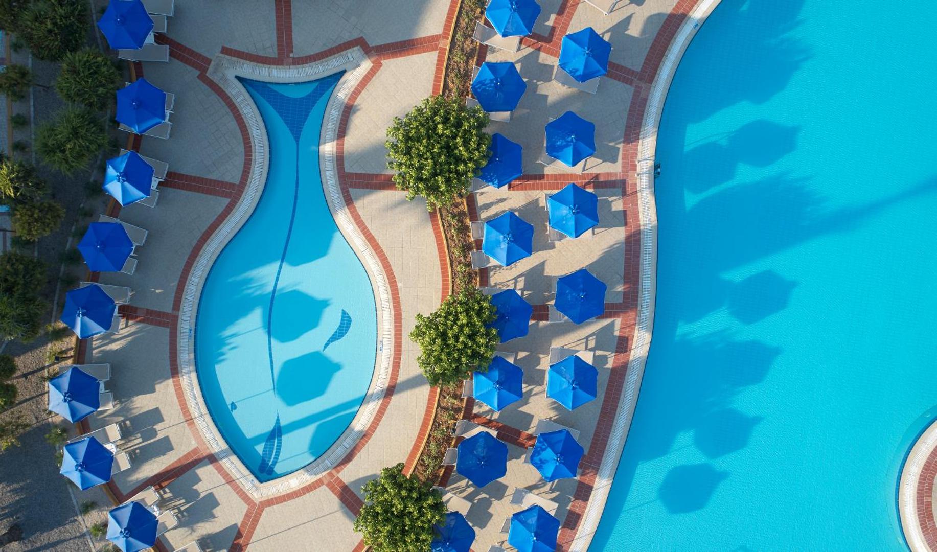 Lindos Princess Beach Hotel skats no augšas uz bērnu baseinu zivtiņas formā — zili saulessargi, atpūtas krēsli un kopti apstādījumi