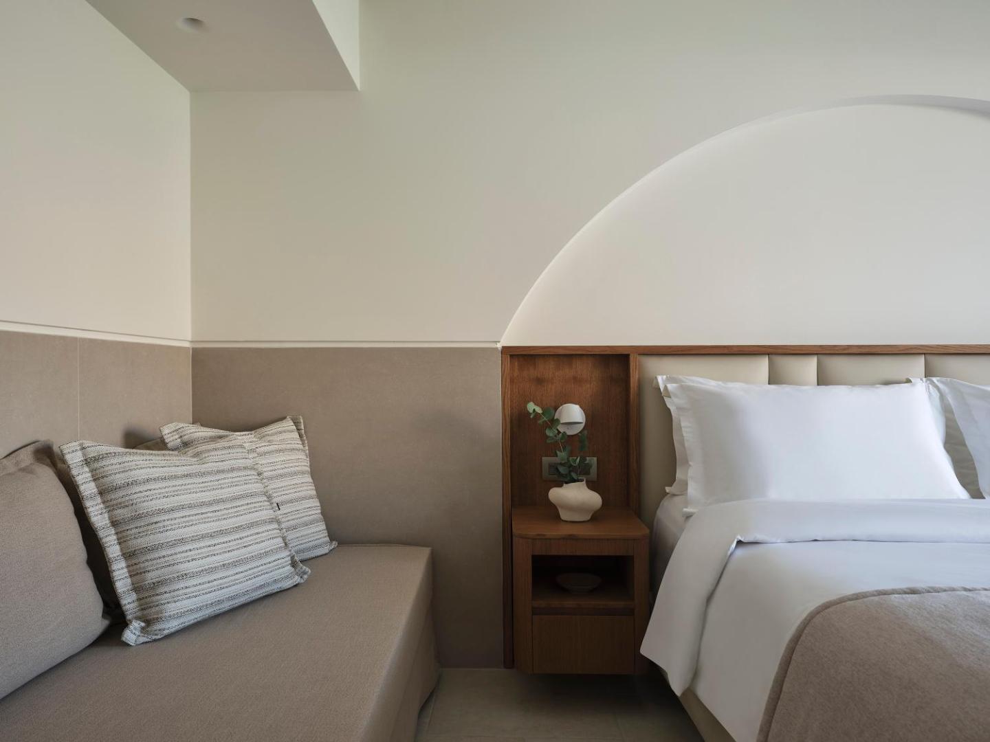 Lindos Princess Beach Hotel numura detaļa ar gultu, naktsgaldiņu un atpūtas dīvānu pie sienas ar dekoratīvu apgaismojumu.