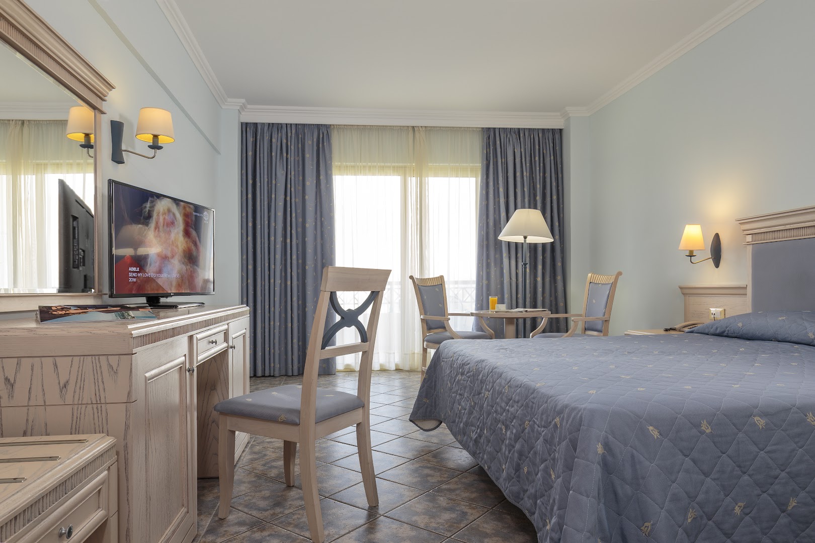 Lindos Princess Beach Hotel klasiskā stila numurs ar zilām detaļām, flīžu grīdu, koka mēbelēm un balkonu Rodas kūrortā.