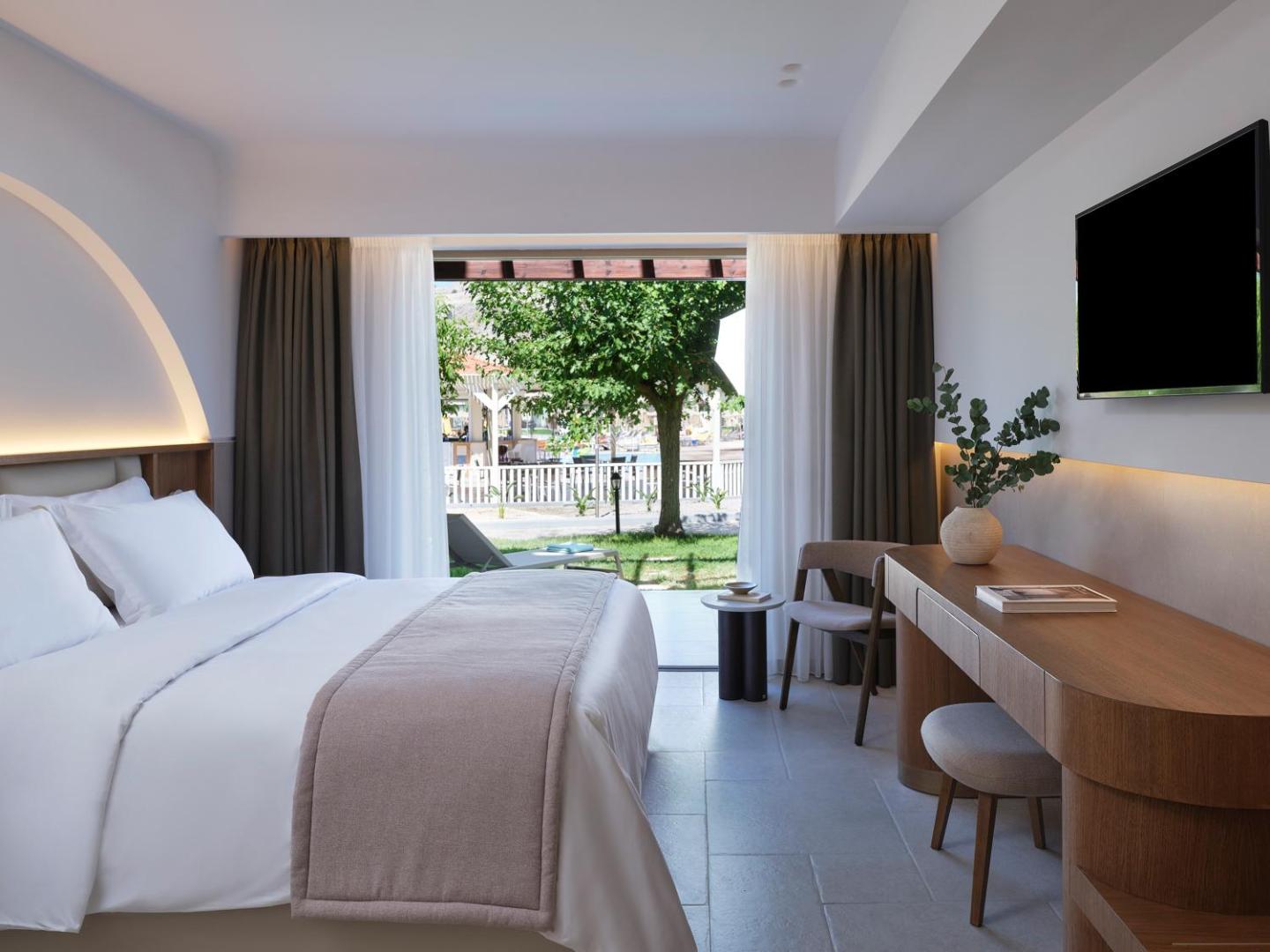 Lindos Princess Beach Hotel numura skats uz dārzu caur atvērtām terases durvīm, gaiša guļamistaba ar rakstāmgaldu un TV.