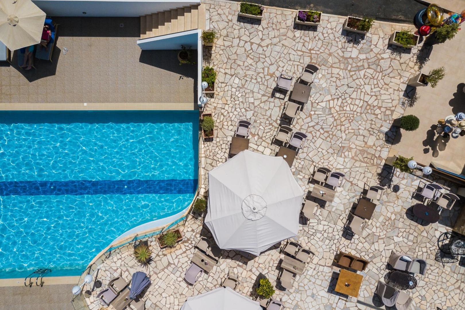 Aerofoto skats uz Filion Suites Resort & Spa baseina terasi ar galdiņiem, saulessargiem un akmens flīžu klājumu no augšas