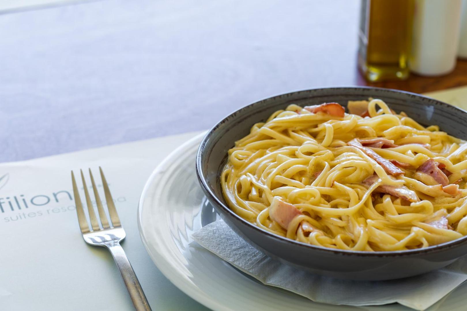 Filion Suites Resort & Spa restorāna piedāvājums — klasiska pasta karbonāra bļodā uz galda ar restorāna logo salveti