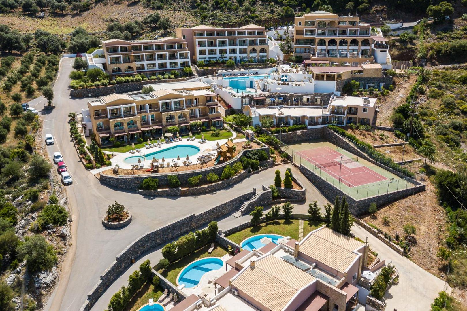 Filion Suites Resort & Spa panorāmas skats no augšas uz baseiniem, tenisa kortu un viesnīcas korpusiem Bali ciematā, Krētā
