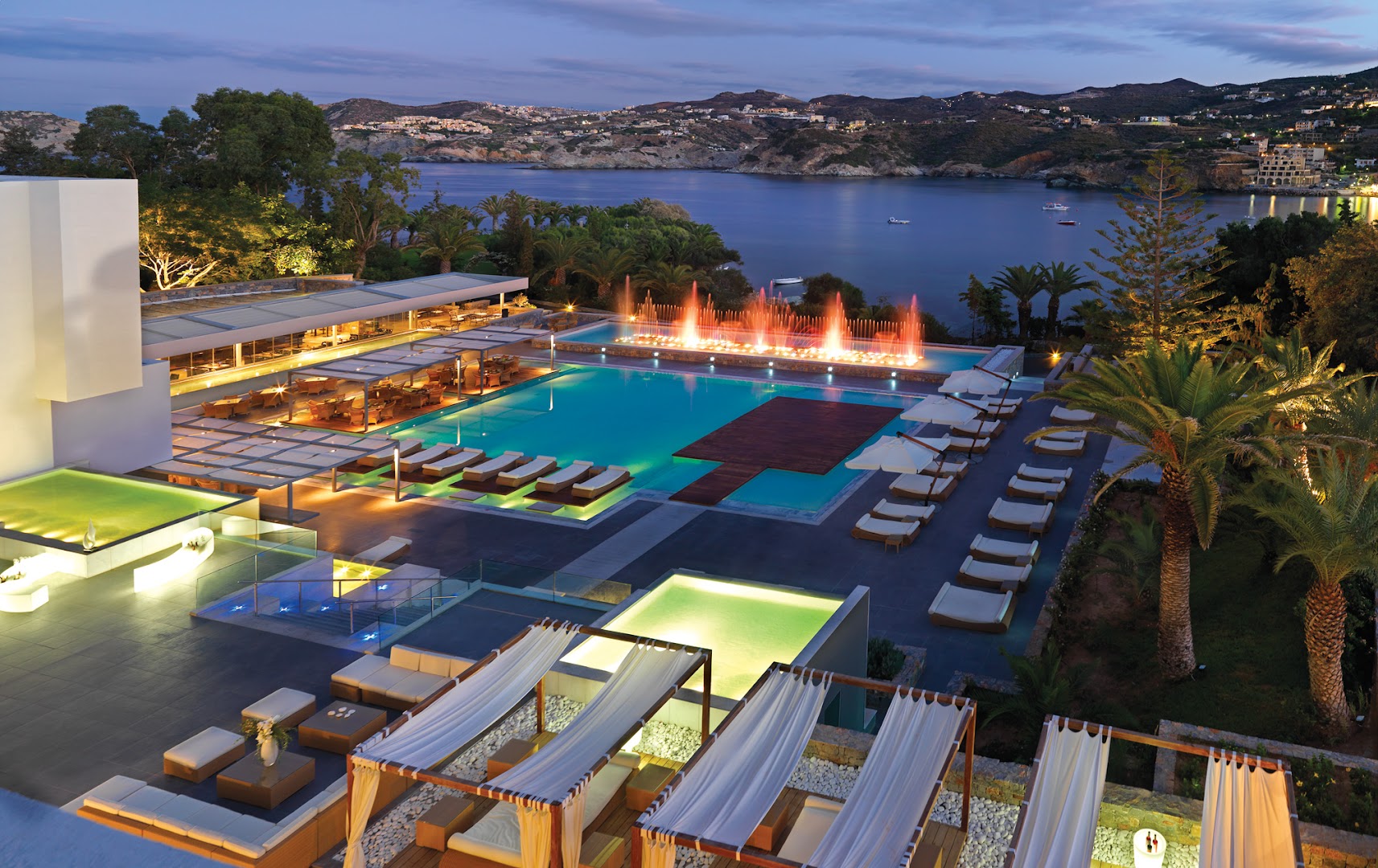 Out Of The Blue Resort & Spa galvenais baseinu komplekss vakarā ar izgaismotām strūklakām un skatu uz Agia Pelagia līci
