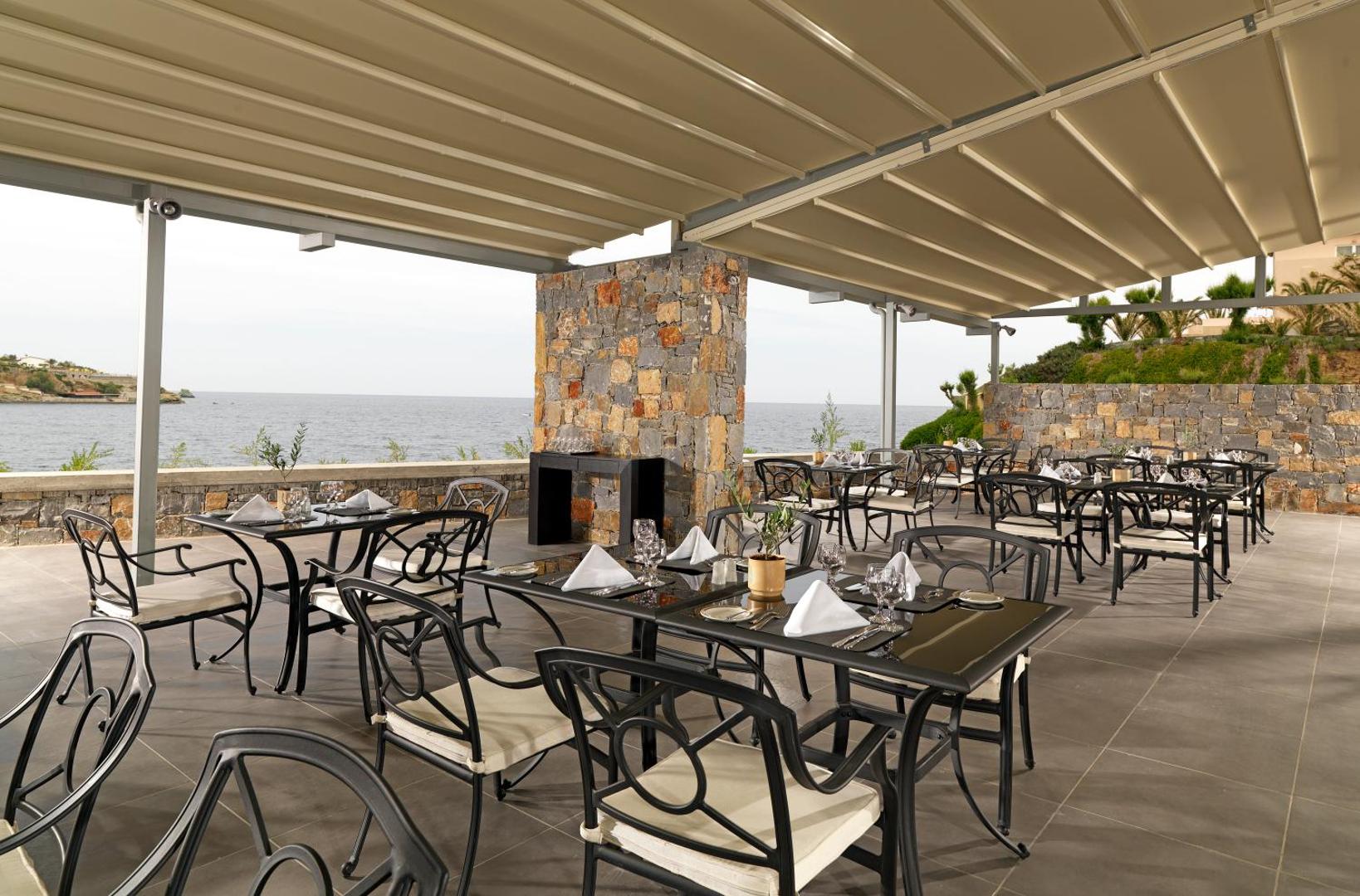 Out Of The Blue Resort & Spa āra terases restorāns ar skatu uz jūru Krētā — eleganti galdiņi zem nojumes pie akmens mūra