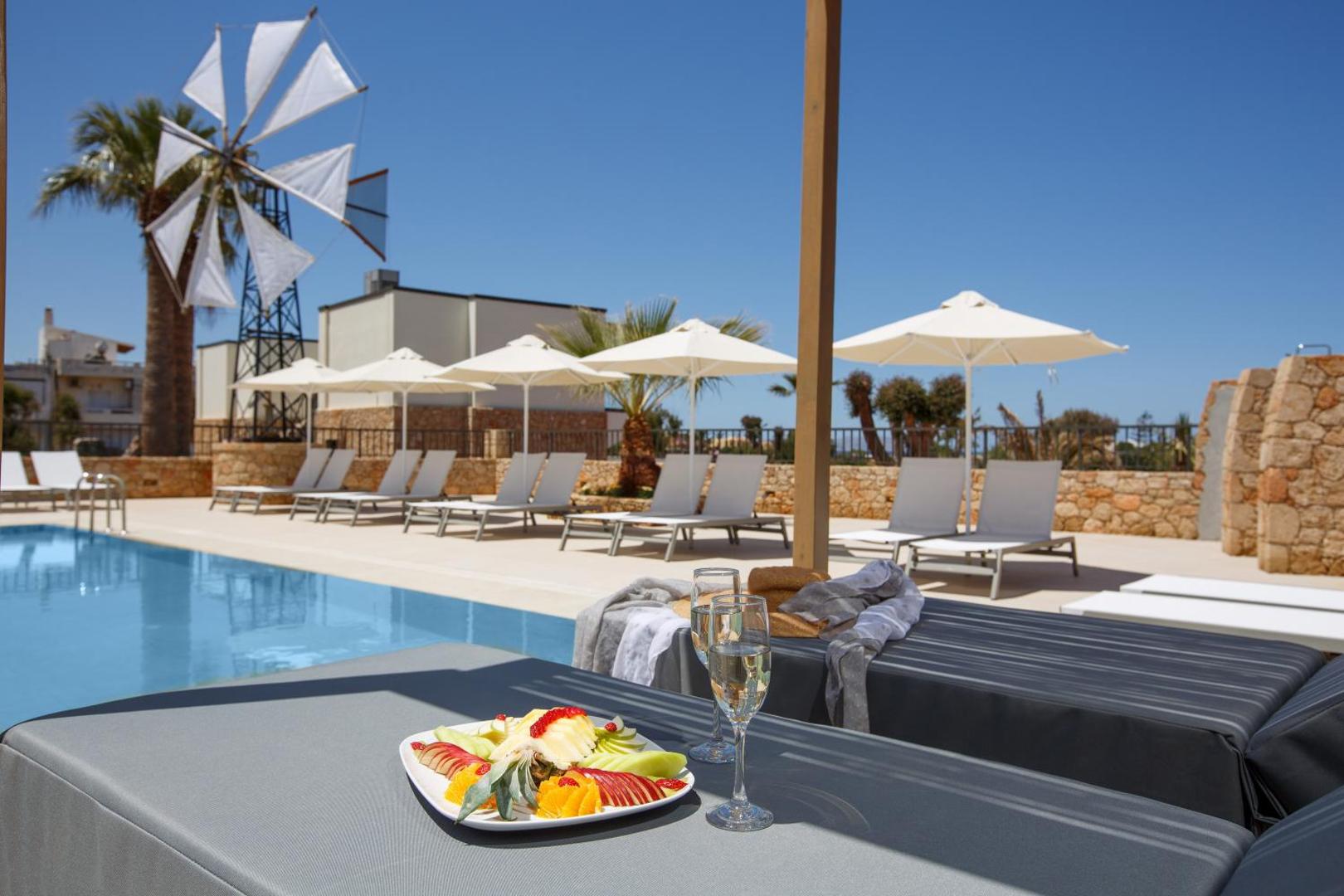 Aloe Boutique Hotel atpūtas zona pie baseina ar augļu plati, dzērieniem, saulessargiem un vējdzirnavu fonā saulainā dienā