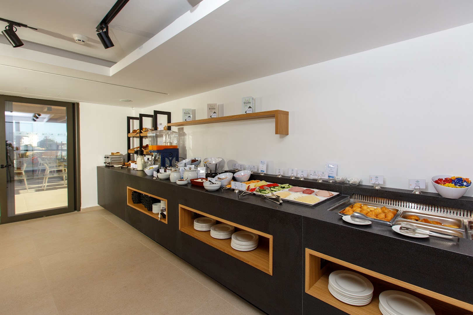 Aloe Boutique Hotel brokastu bufetes zona ar plašu uzkodu, sieru un augļu izvēli modernā un gaišā interjerā