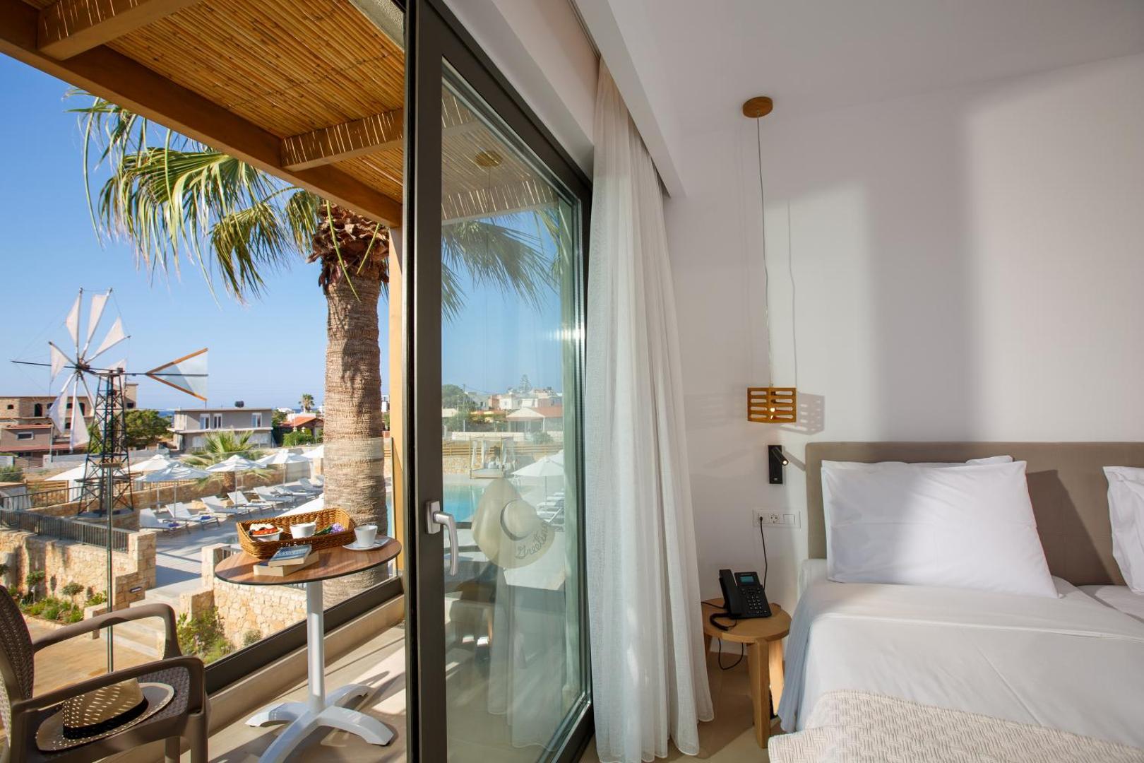 Aloe Boutique Hotel numurs ar balkonu, skatu uz baseinu un tradicionālu vējdzirnavu Hersonisā, Krētā — brokastu galdiņš un palmas.