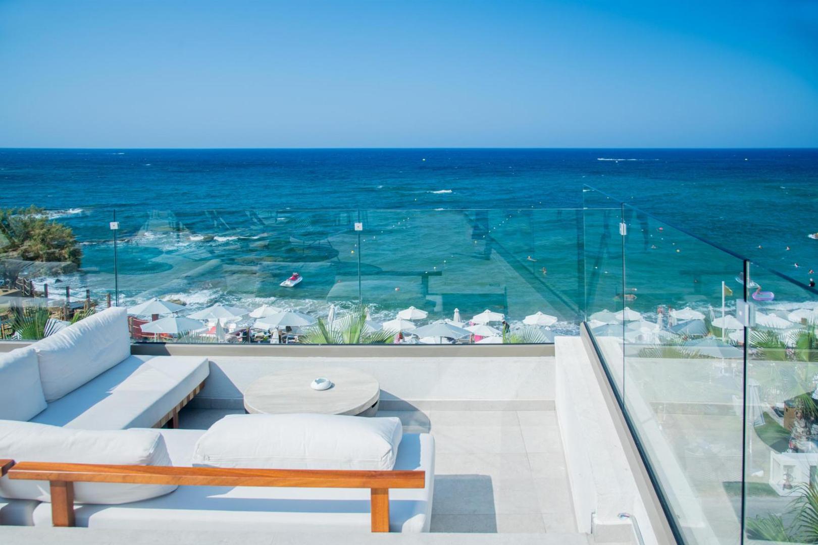 Skats no Akasha Beach Hotel & Spa terases ar stikla margām uz zilo Vidusjūru — balti dīvāni atpūtai un pludmales panorāma