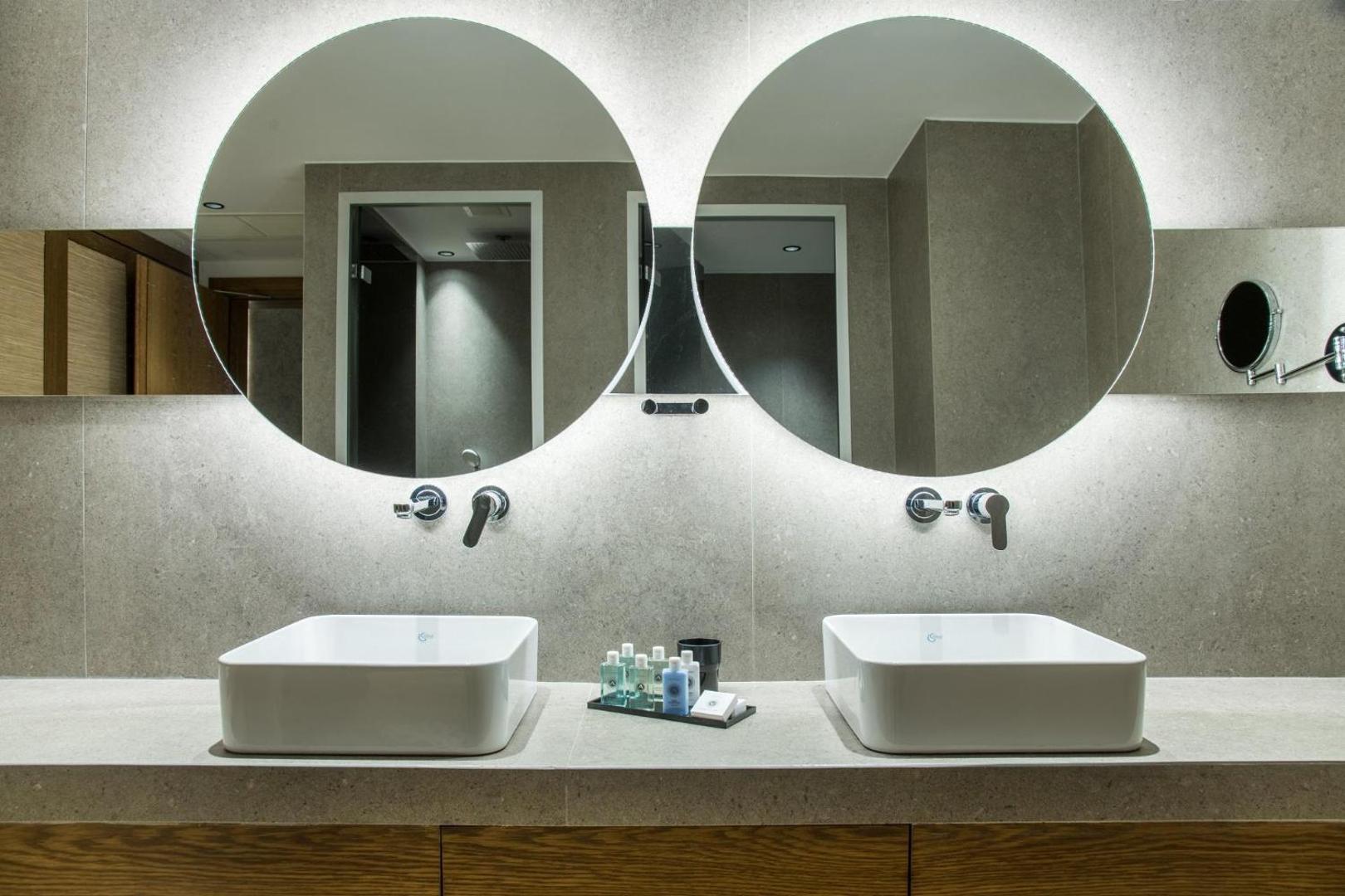 Akasha Beach Hotel & Spa moderna vannas istaba ar divām izlietnēm, apaļiem spoguļiem un apgaismojumu — elegants dizains Krētas kūrortā