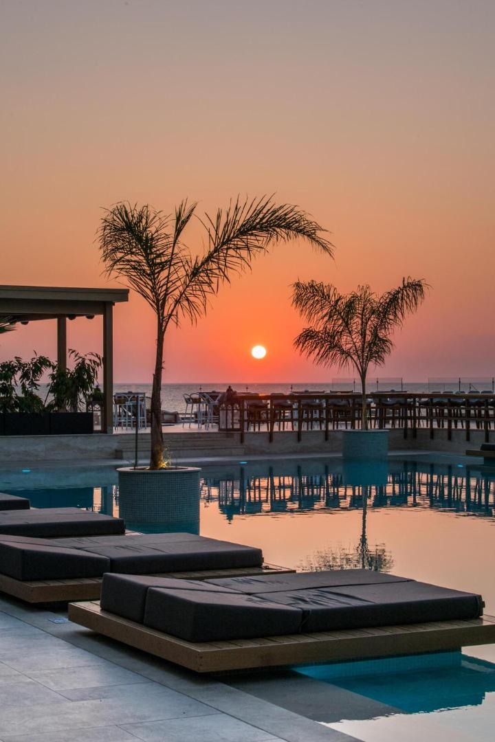 Saulriets virs Vidusjūras pie Akasha Beach Hotel & Spa — palmu silueti un atpūtas gultas pie baseina malas