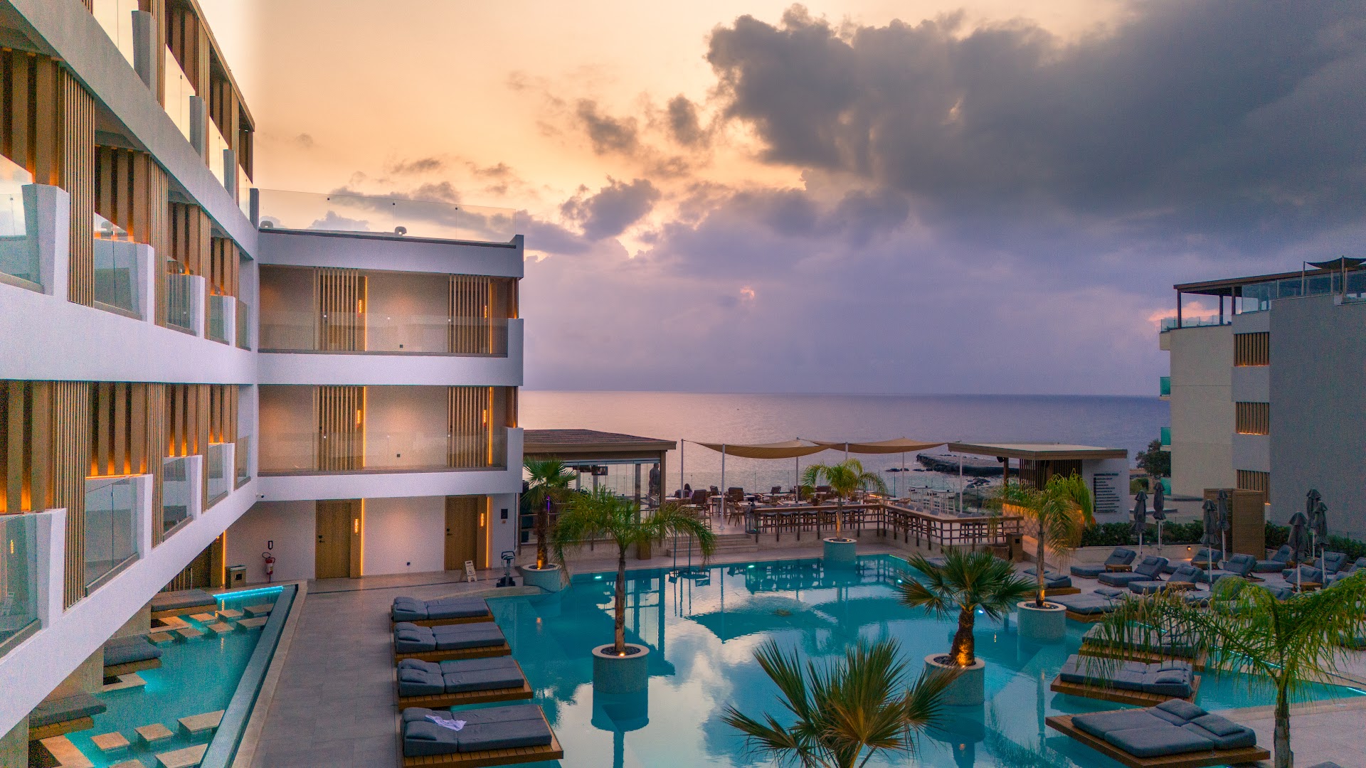 Akasha Beach Hotel & Spa baseina zona saulrietā ar skatu uz jūru — moderna viesnīcas fasāde un palmas piekrastē