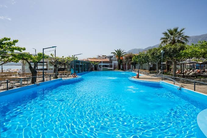 Corissia Princess plašais āra baseins ar dzidru ūdeni, ko ieskauj palmas un atpūtas zona Krētas kalnu fonā.