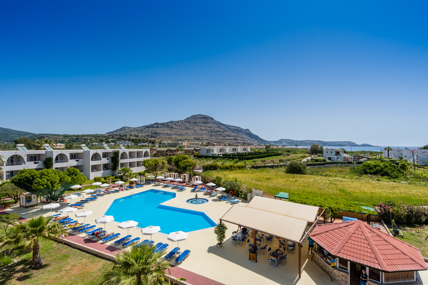 Lardos Bay viesnīcas kopskats Rodas salā ar āra baseinu, atpūtas krēsliem un kalnu fonā saulainā dienā
