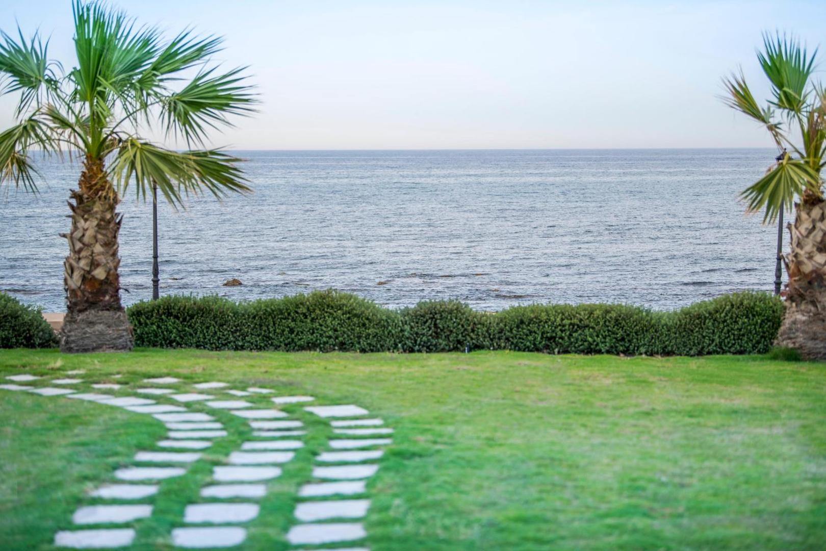 Skats uz Vidusjūru no Insula Alba Resort & Spa dārza — zaļš zālājs ar akmens taciņu un palmām piekrastē