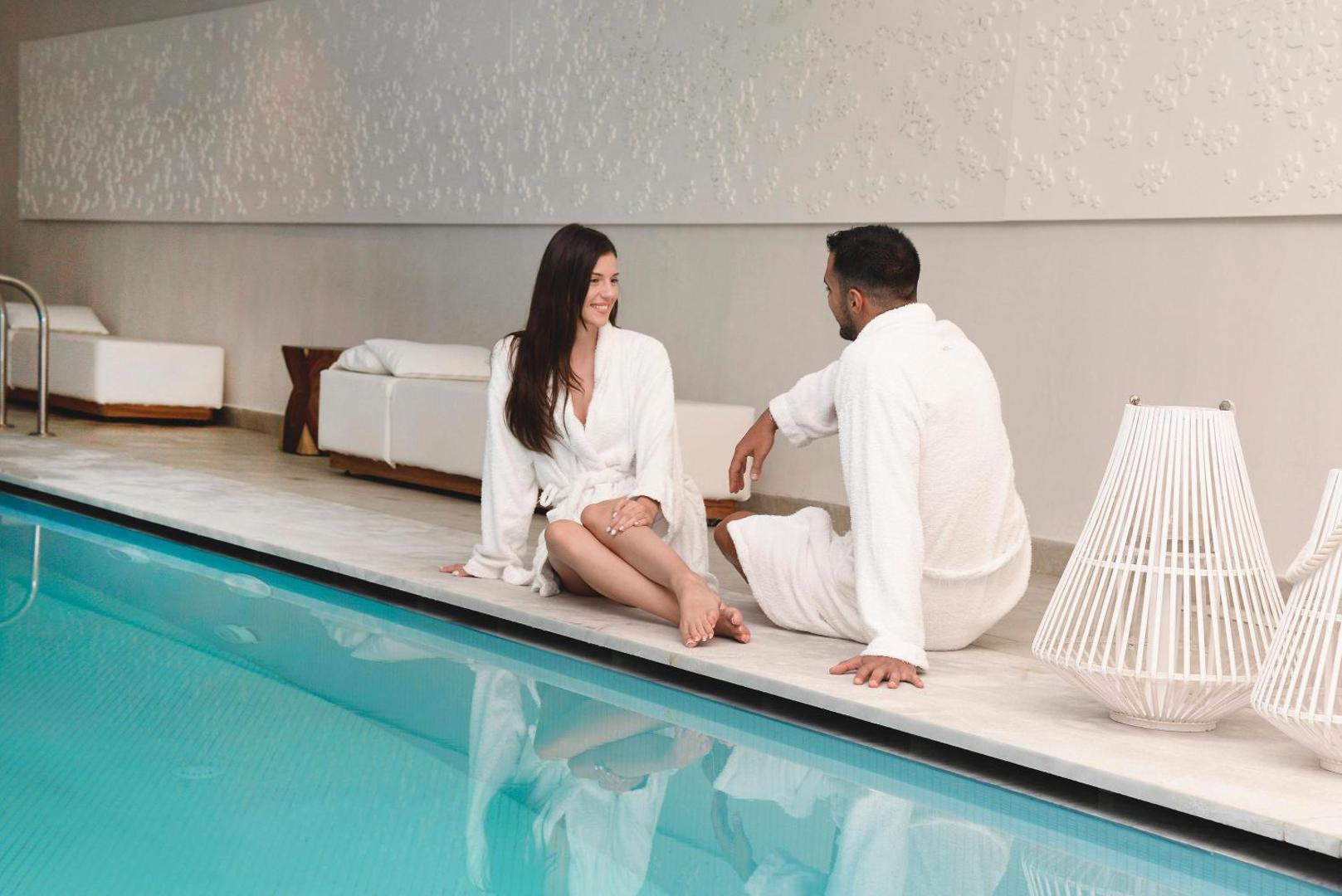 Pāris baltos peldmēteļos atpūšas pie Insula Alba Resort & Spa iekštelpu baseina modernajā spa centrā Krētā