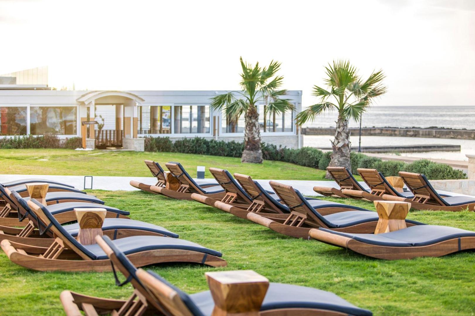 Eleganti koka sauļošanās krēsli Insula Alba Resort & Spa teritorijā ar skatu uz viesnīcas ēku un palmām