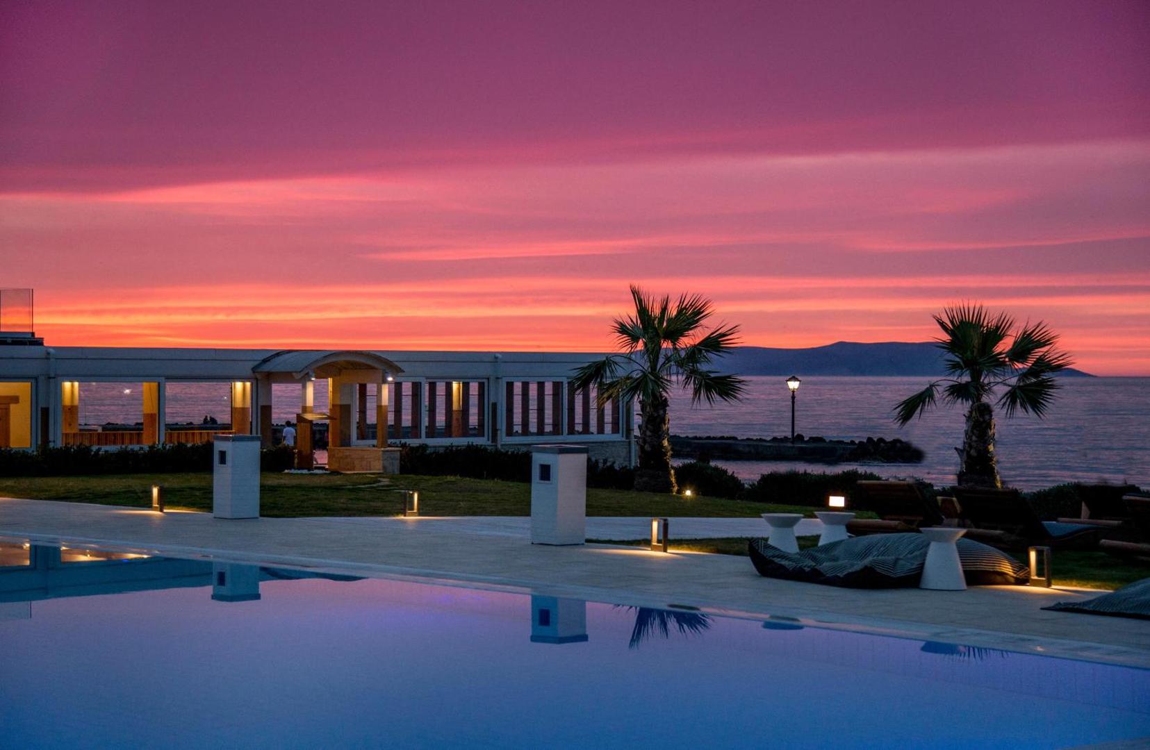 Insula Alba Resort & Spa baseins saulrieta laikā ar skatu uz jūru — rozā debesis, palmas un relaksācijas zona piekrastē