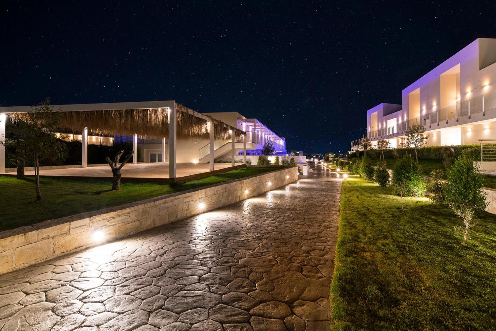 Insula Alba Resort & Spa izgaismota teritorija naktī ar akmens flīžu celiņu, modernām ēkām un zvaigžņotām debesīm Hersonisā, Krētā.