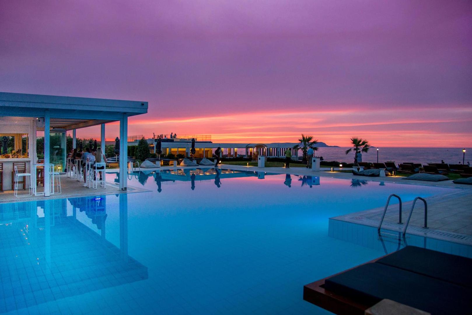 Insula Alba Resort & Spa āra baseins saulrietā Krētā — baseina bārs ar apgaismojumu un rozā debesu atspulgu ūdenī