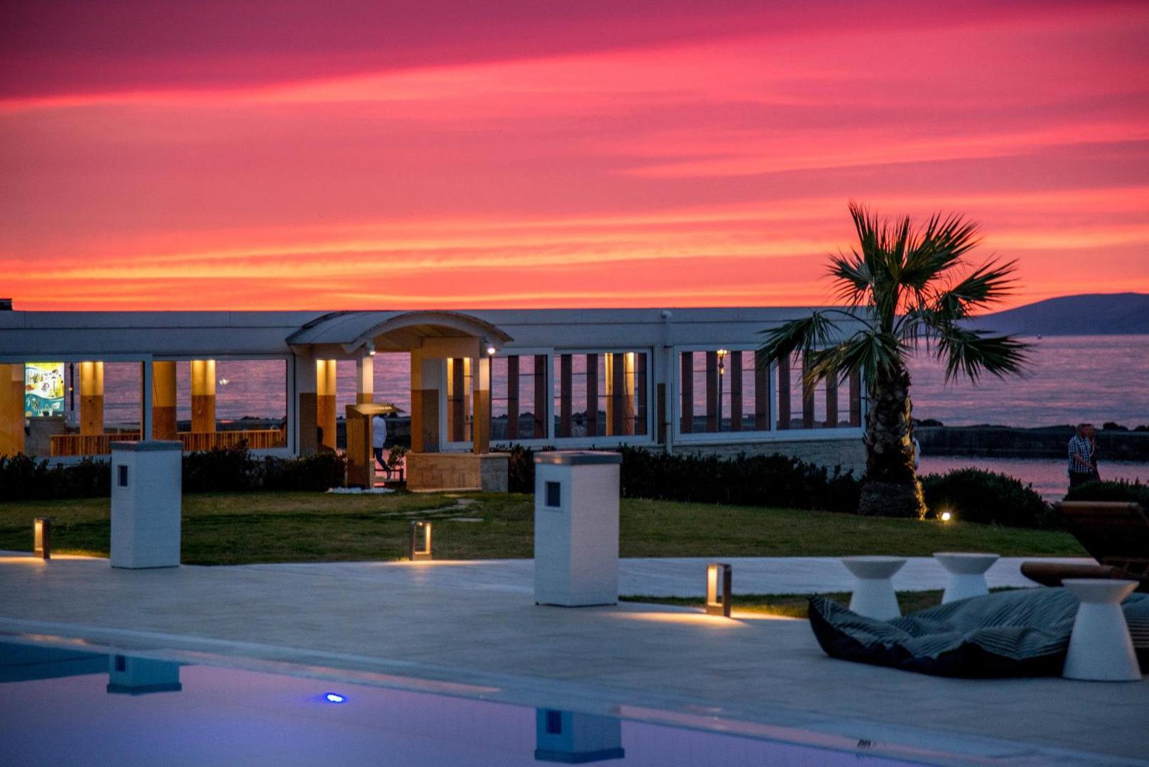 Saulriets Insula Alba Resort & Spa piekrastē ar rozā debesīm, baseina malu priekšplānā un palmu siluetiem Krētas salā.