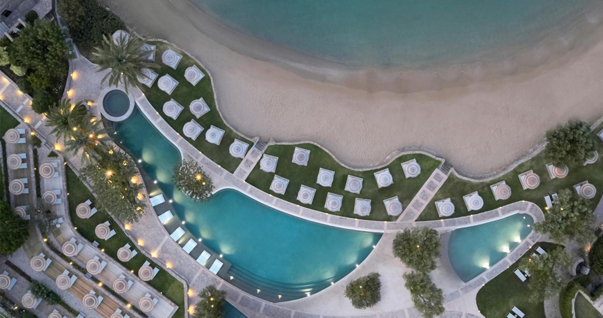 Porto Elounda Golf & Spa Resort aerofoto ar izliektas formas baseinu, saulessargiem un smilšu pludmali pie jūras