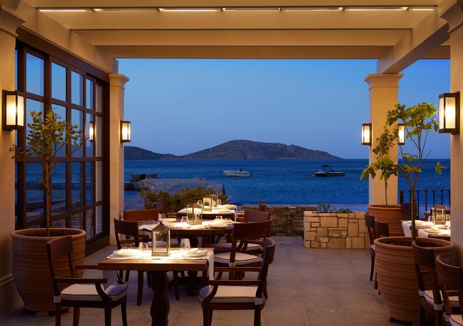 Porto Elounda Golf & Spa Resort ēdienkartes restorāna terase vakarā ar galdiņu klājumu un skatu uz jūru un kalniem