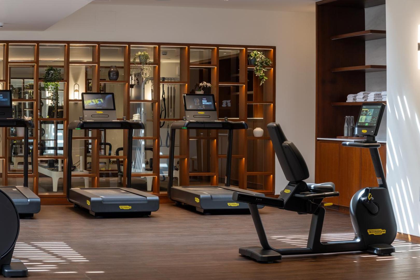 Porto Elounda Golf & Spa Resort modernā fitnesa zāle ar Technogym skrejceļiem un velotrenažieri — gaišs interjers ar koka apdari