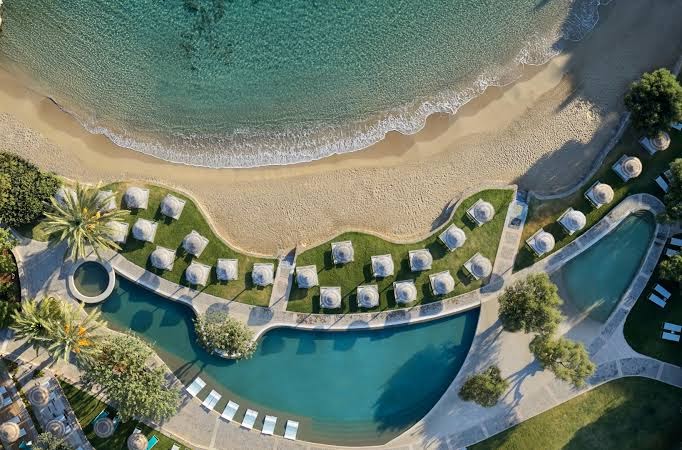 Porto Elounda Golf & Spa Resort teritorijas skats no augšas uz baseinu, koptu zālienu un pieguļošo jūras līci