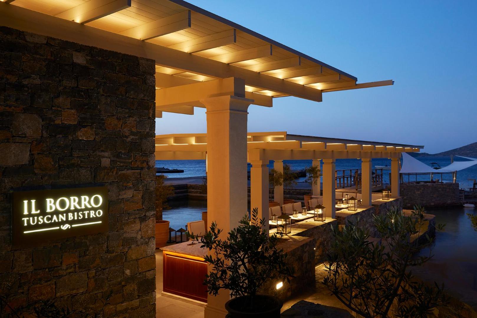 Il Borro Tuscan Bistro ieeja Porto Elounda kūrortā vakarā — akmens sienas, silts apgaismojums un terase pie jūras