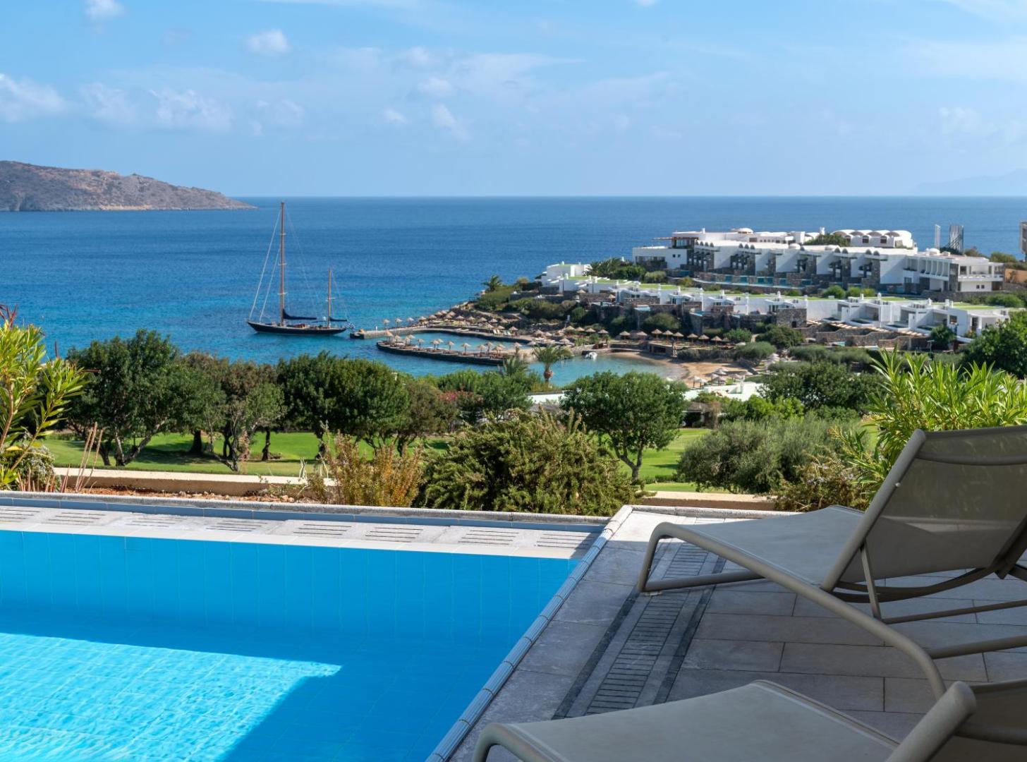Skats no Porto Elounda Golf & Spa Resort terases uz kūrorta ēkām, jahtu un līci saulainā dienā