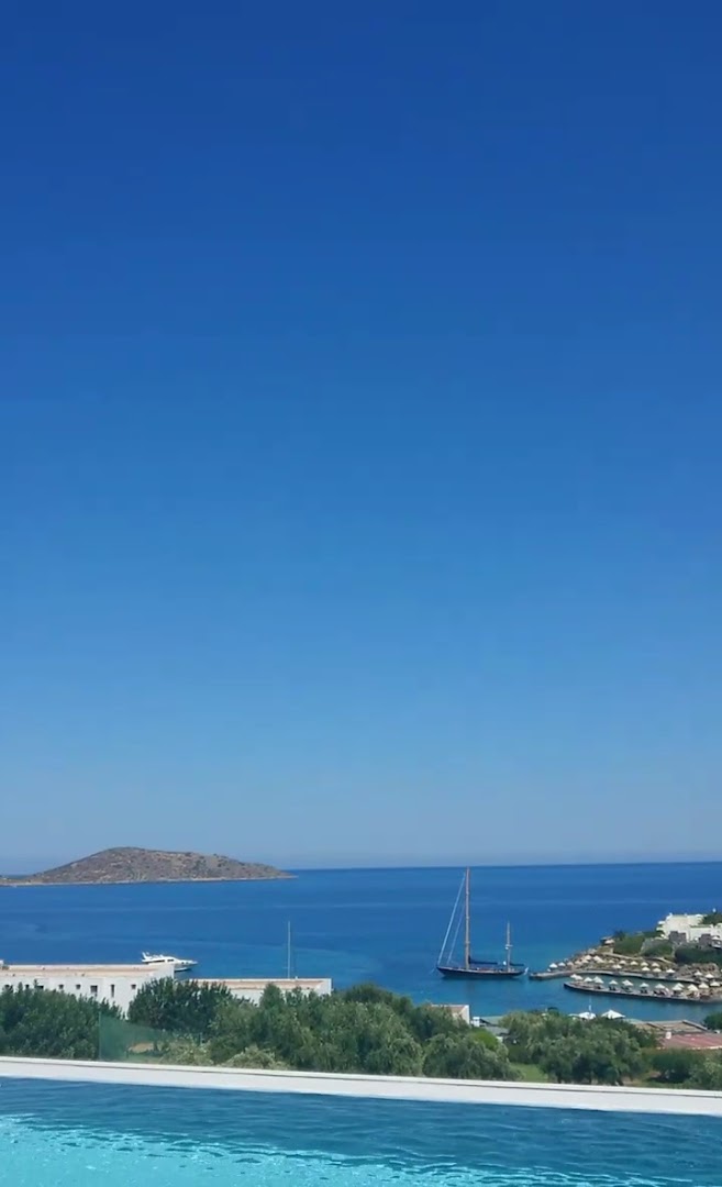 Skats no Porto Elounda Golf & Spa Resort baseina malas uz Mirabello līci Krētā ar jahtām un zilu debesu horizontu